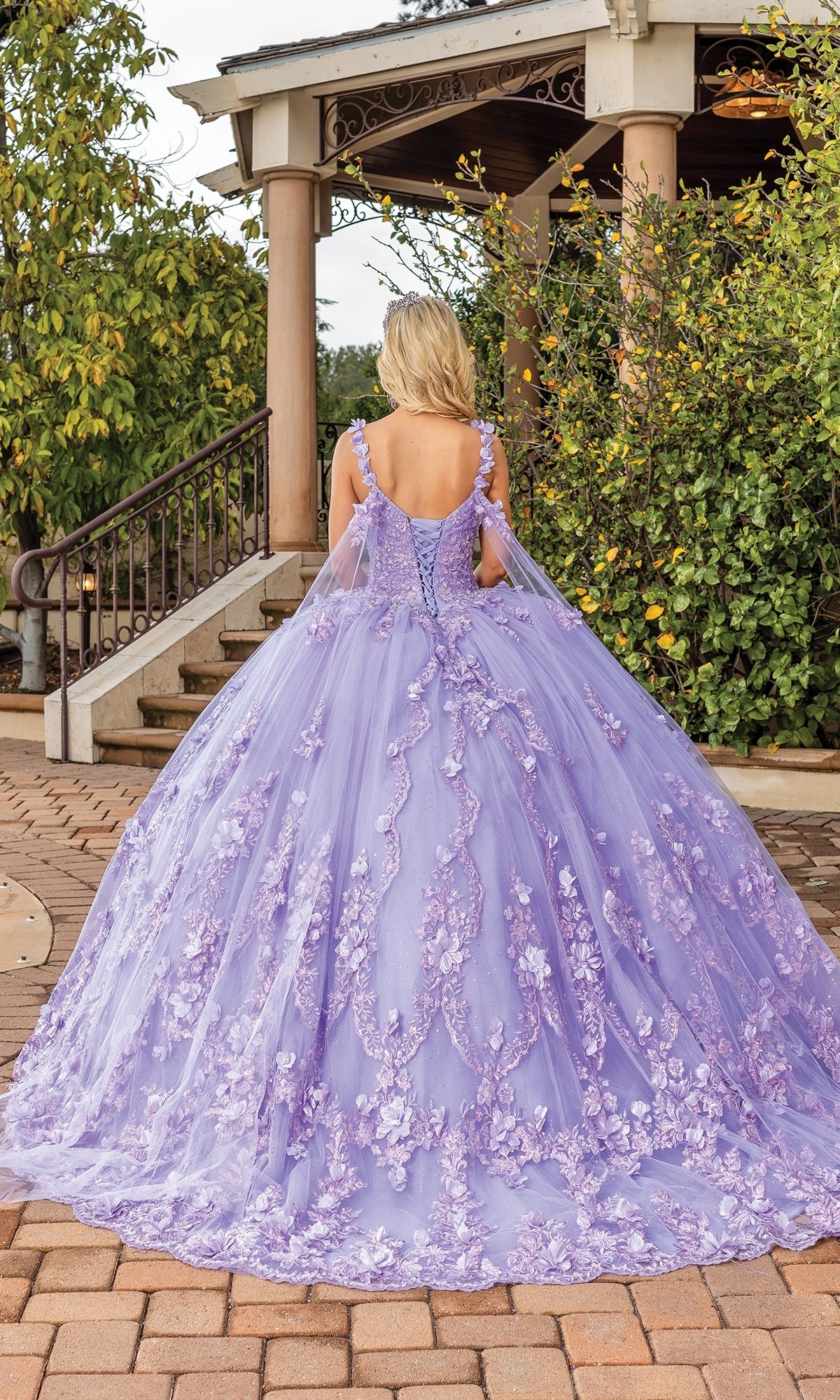 A woman stands outside facing away in front of a gazebo, wearing the Quinceanera Dress 1808 By Dancing Queen—a floor-length lavender ball gown with floral embroidery and lace appliqué.